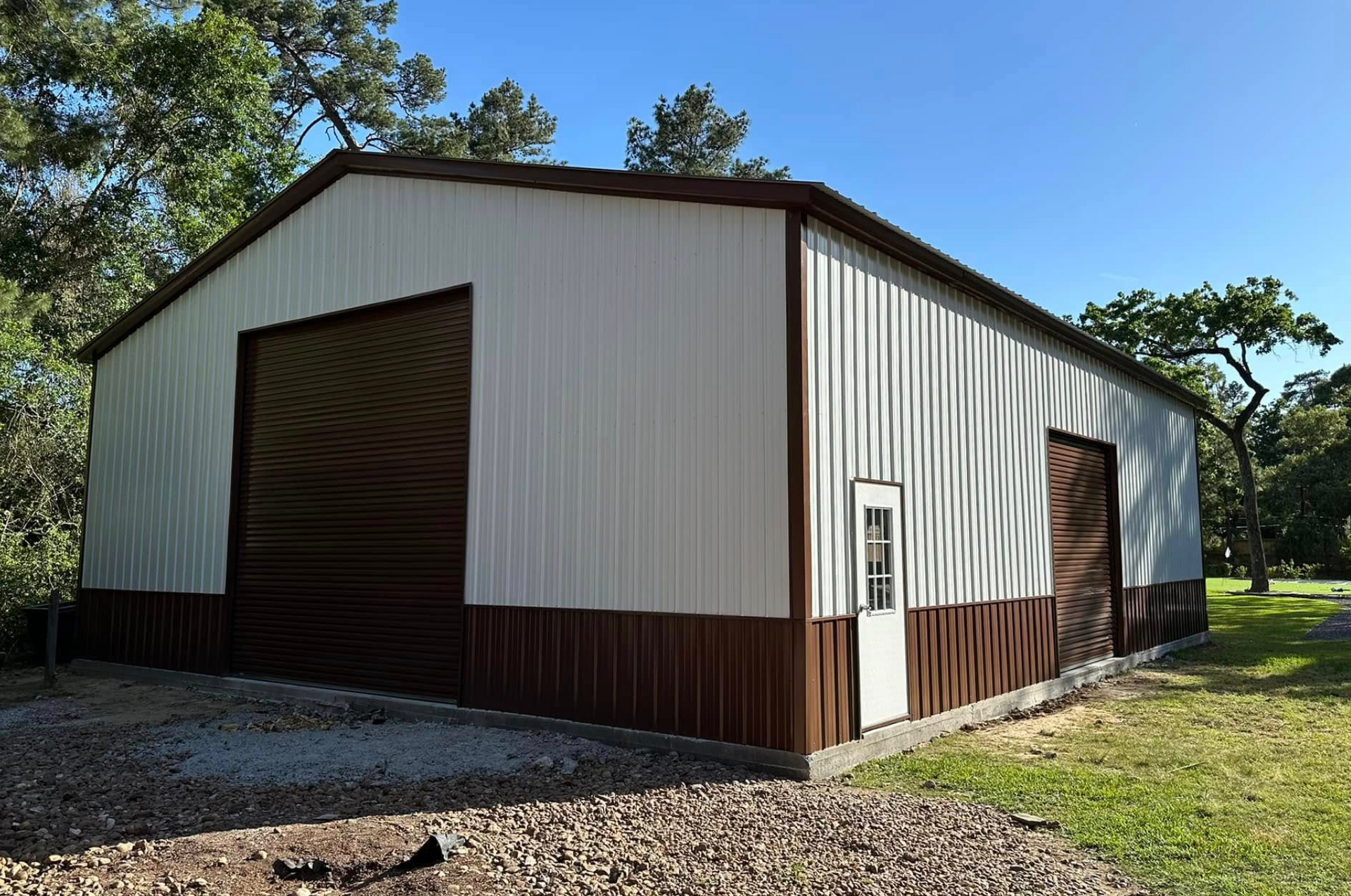 White metal storage building with brown trim on a gravel lot, surrounded by trees under a clear blue sky