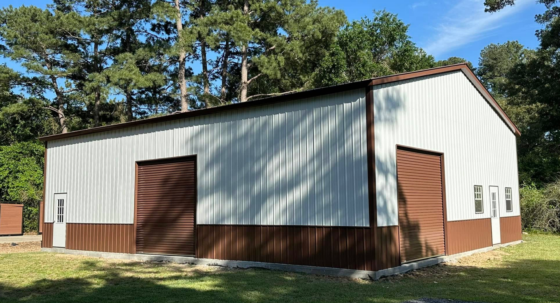 White metal shed with brown trim and doors in a wooded yard under blue sky