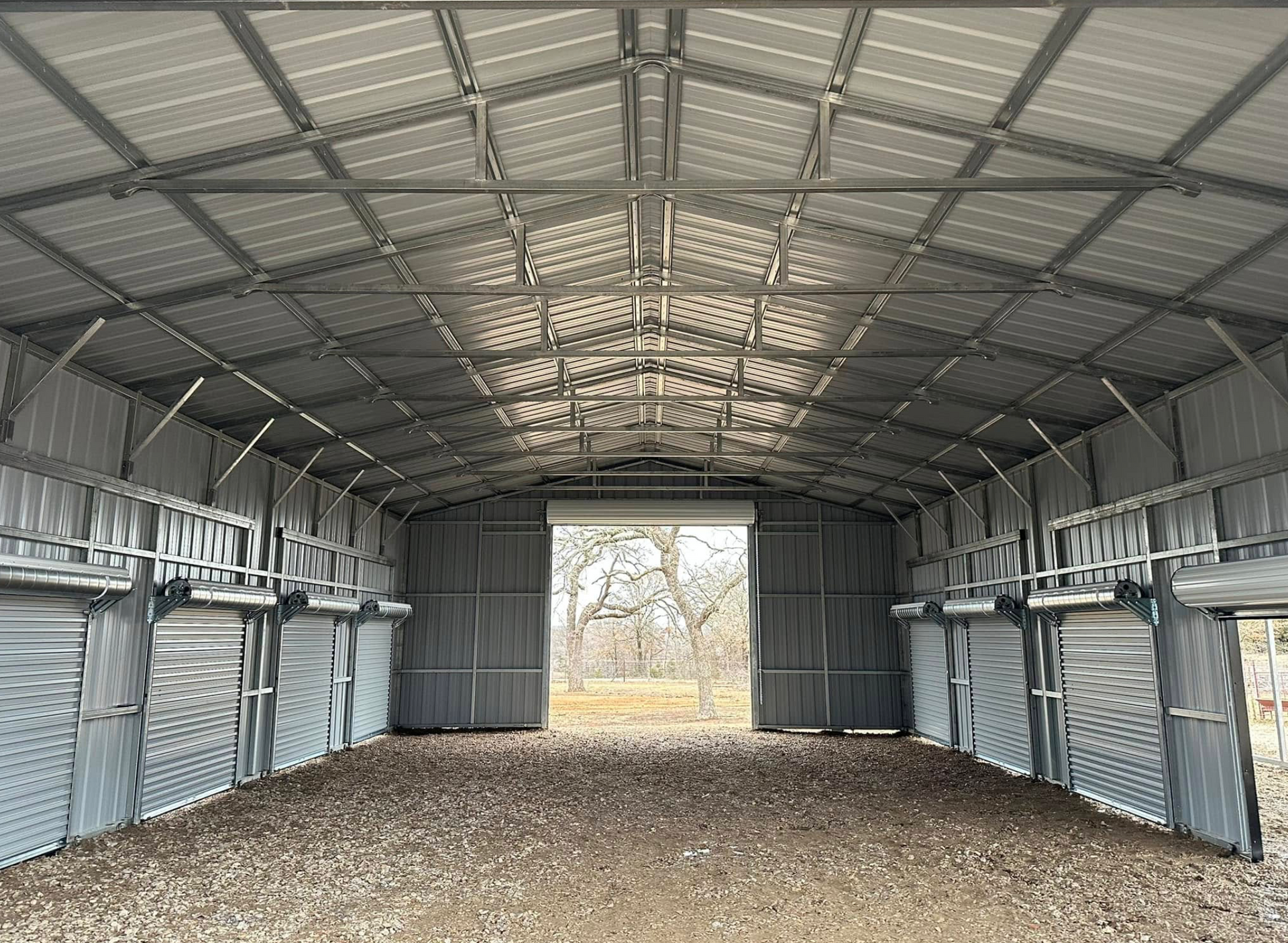 Empty metal shed with corrugated walls, open end, and gravel floor under a high truss roof.