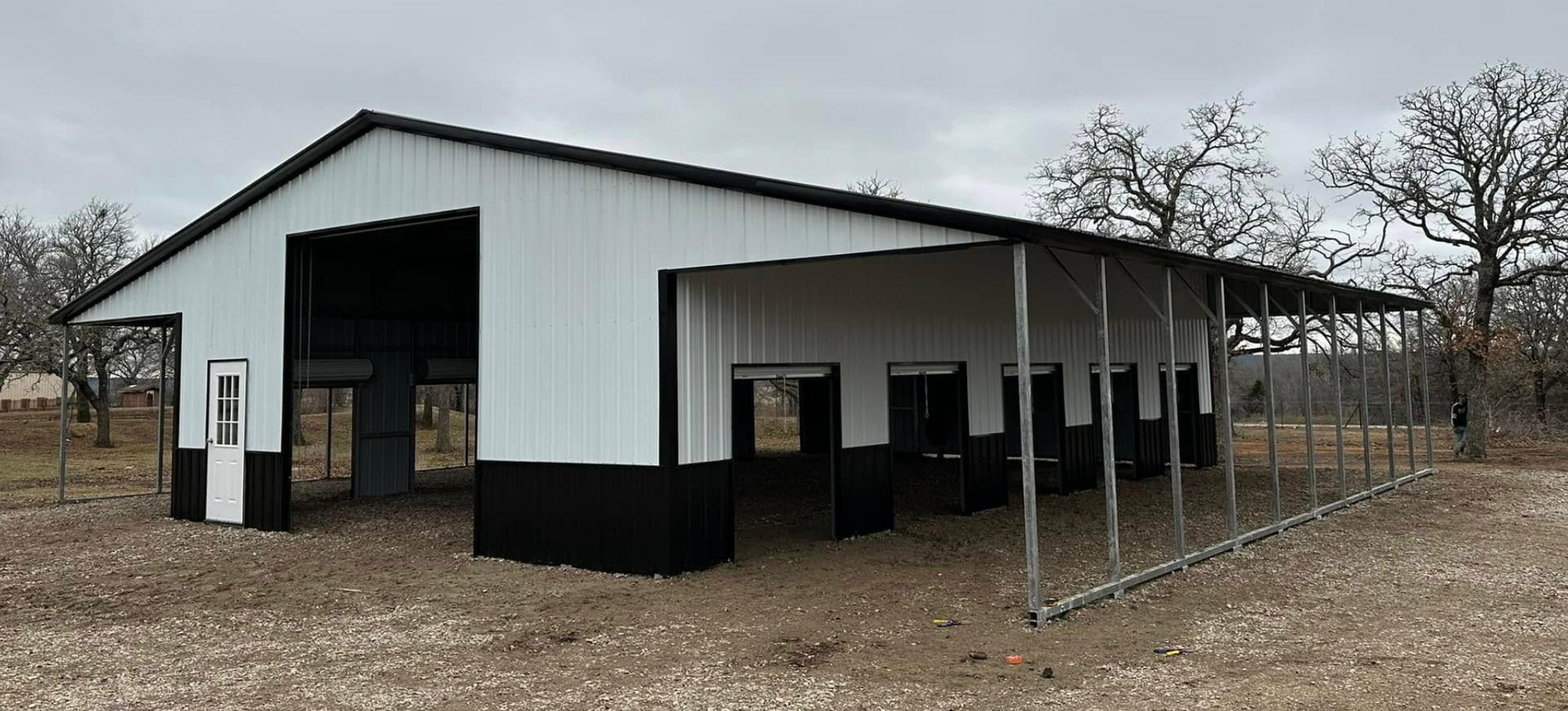 White barn-style shed with black trim and open front on a gravel lot, beside a fenced area.