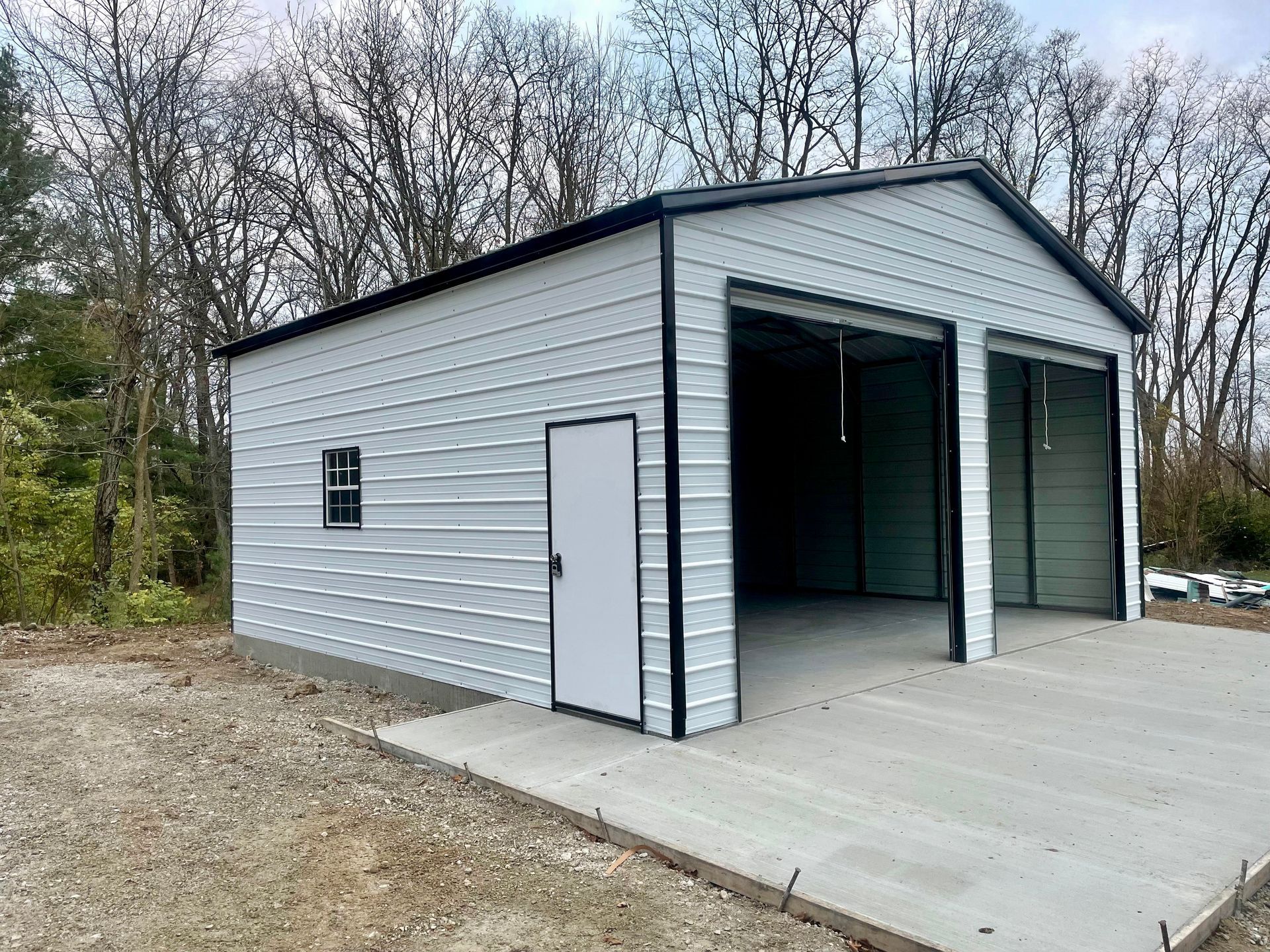 White metal garage with open bays and a side door on a concrete slab, set beside trees.