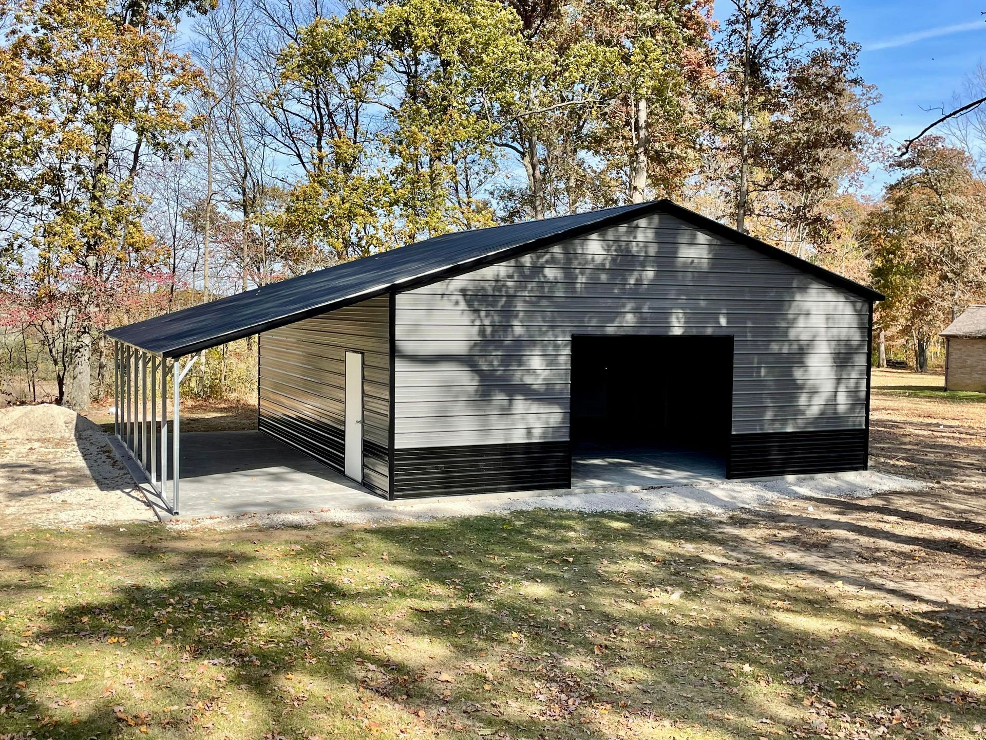 Black metal carport and garage with an open bay in a wooded yard on a sunny day