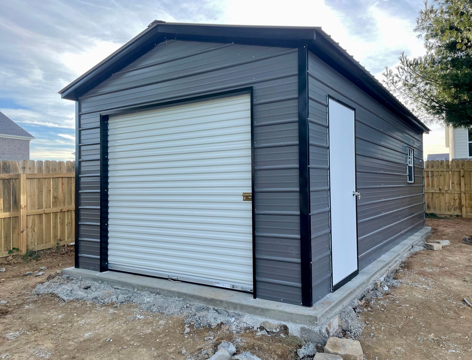 Gray metal garage shed with roll-up door and side door on a fenced dirt lot