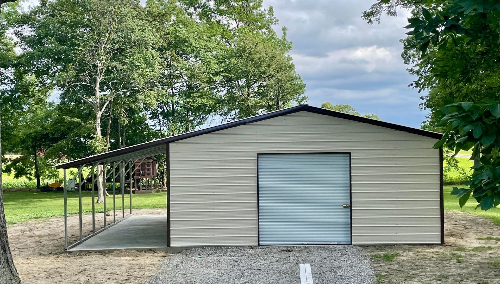 Small beige metal shed with a blue roll-up door on a gravel lot, surrounded by trees under a cloudy sky
