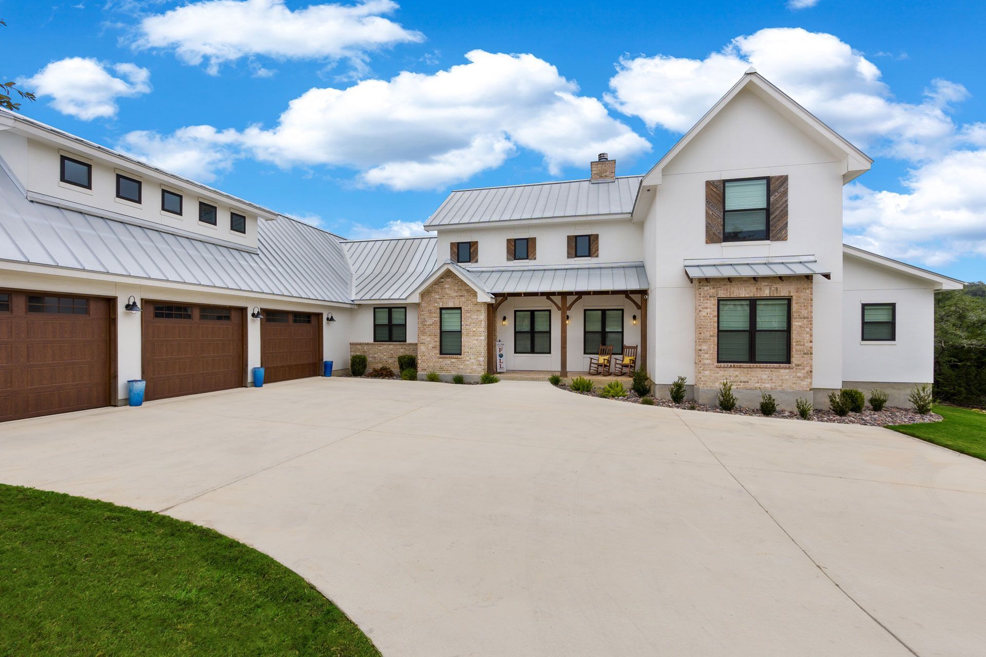 A large, modern white farmhouse with a metal roof, stone accents, brown garage doors, and a concrete driveway.