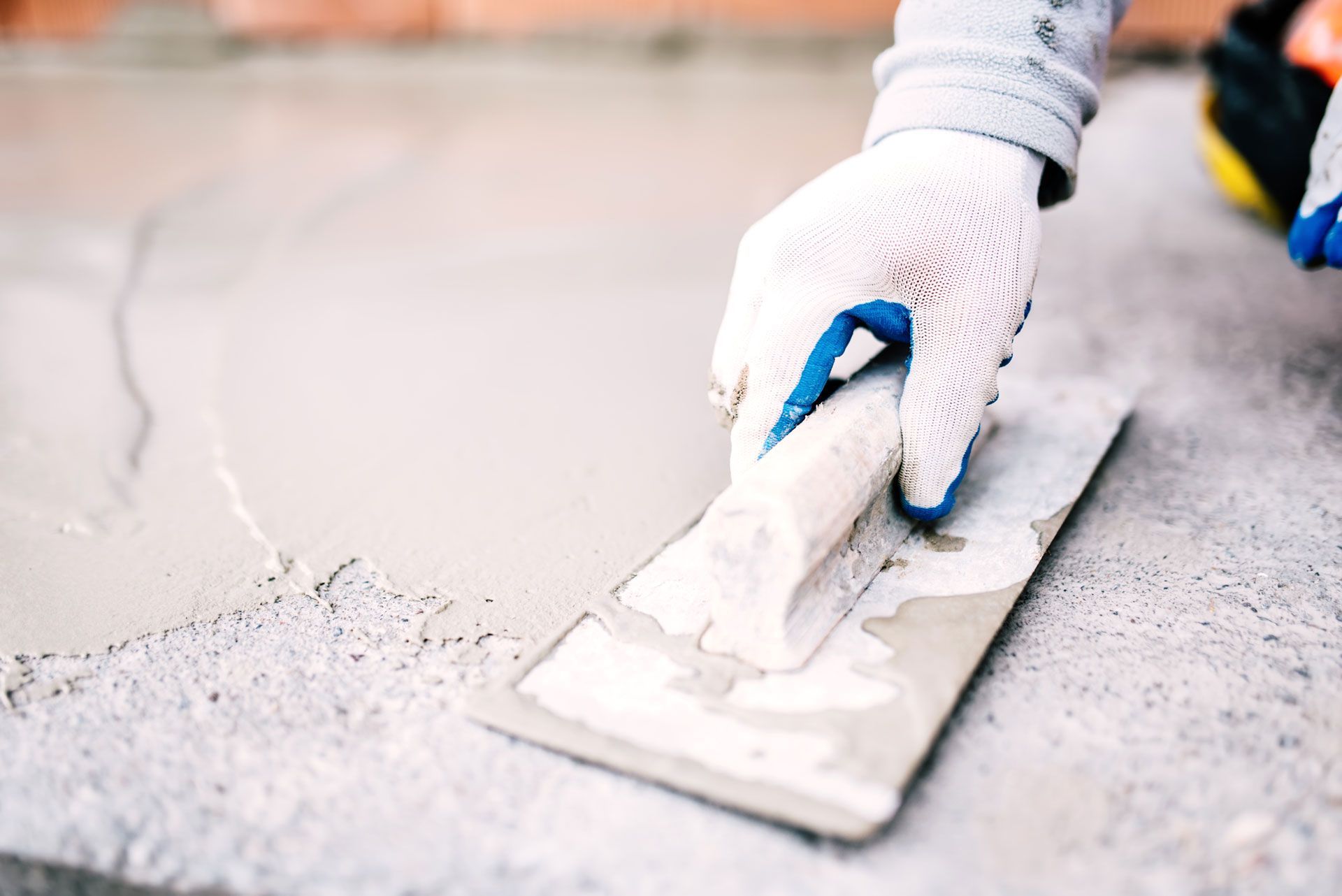 A worker wearing white textured gloves uses a flat trowel to smooth wet concrete onto a floor surface.
