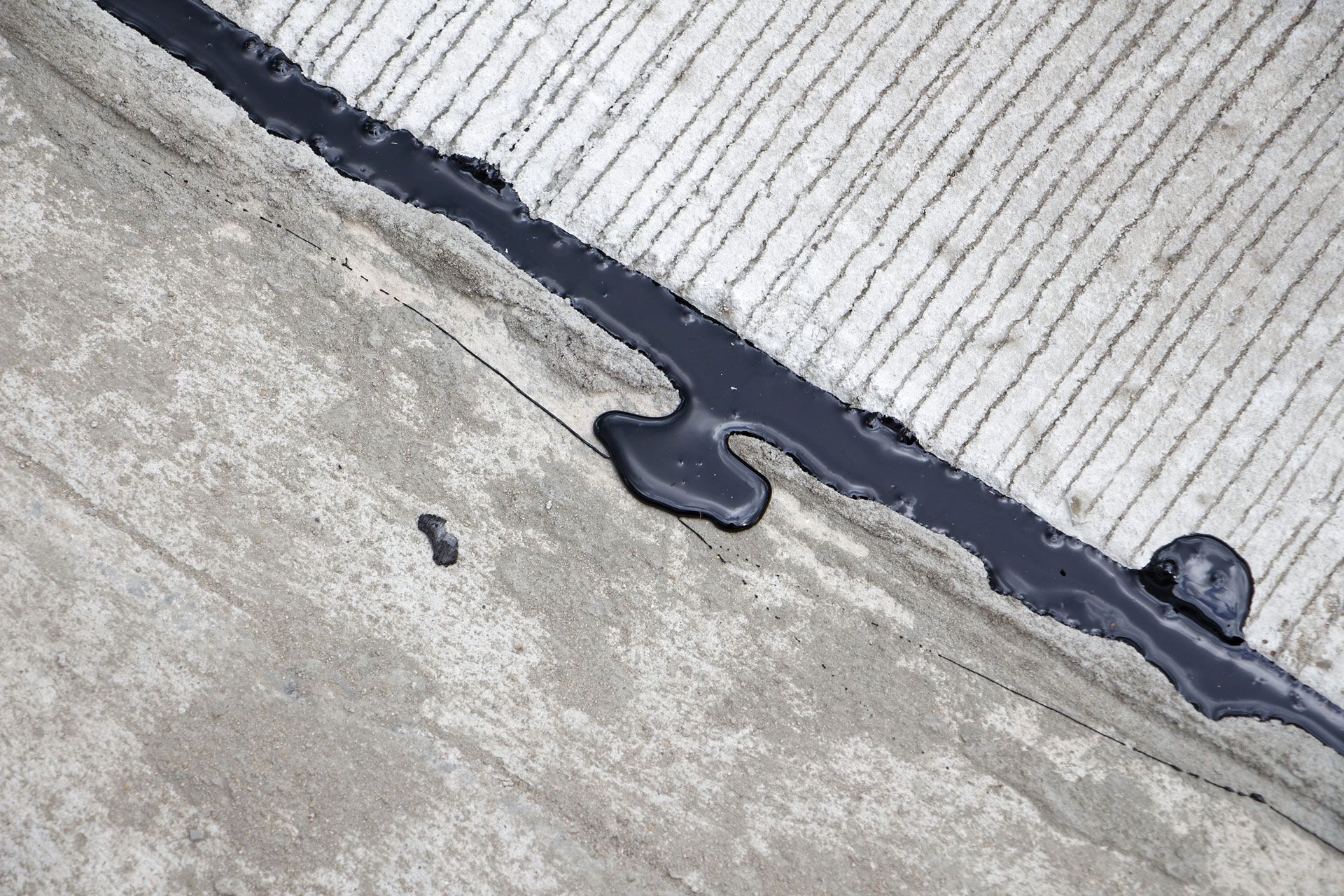 A diagonal crack in a concrete surface filled with dark black sealant, spilling slightly onto the textured concrete.