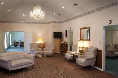 Elegant hotel room with cream chairs, chandelier, lamps, mirrors, and open doorway to a bedroom.