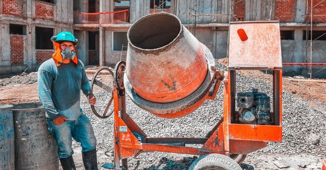 Obrero de la construcción junto a una hormigonera en una obra, con un chaleco de seguridad verde azulado y un casco naranja.