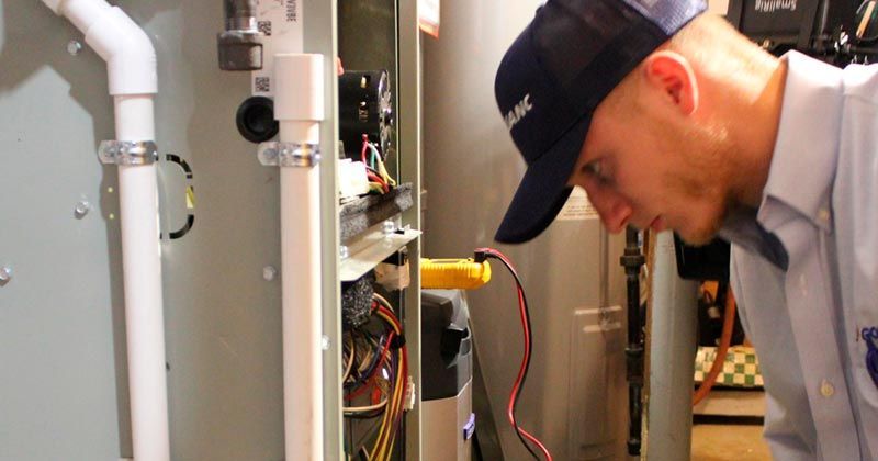 Technician inspecting open electrical panel in a utility room, with exposed wiring and pipes nearby