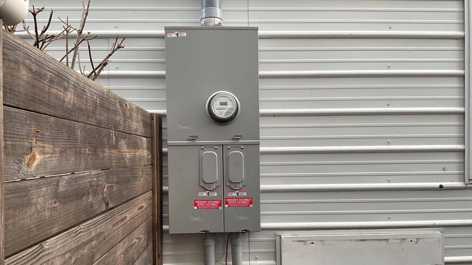 A gray electrical meter and service panel mounted on a metal-sided wall next to a wooden fence.