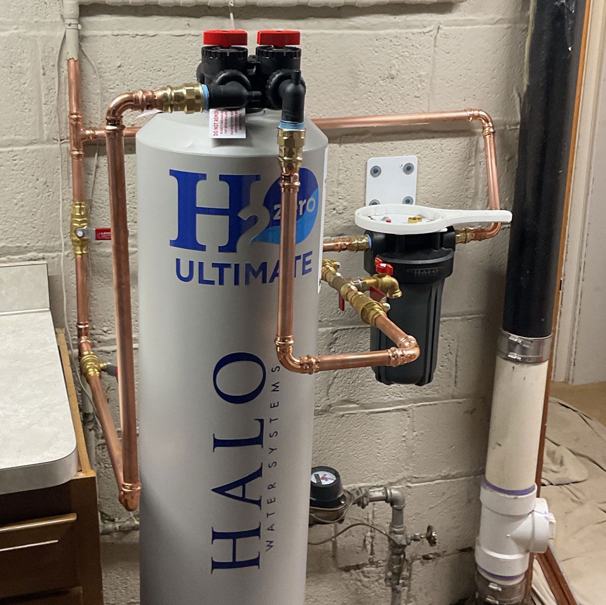 A gray Halo water filtration tank connected by copper pipes to a wall-mounted sediment filter in a basement.