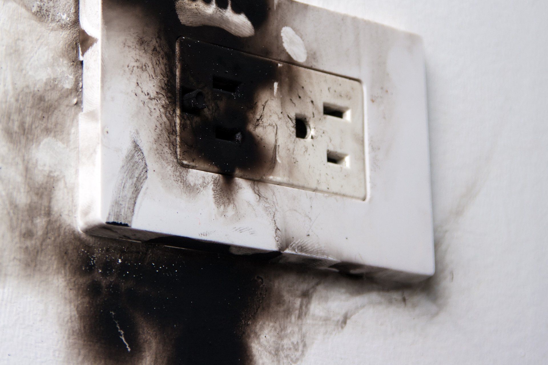 A white electrical wall outlet with significant burn damage and soot staining the surrounding wall.