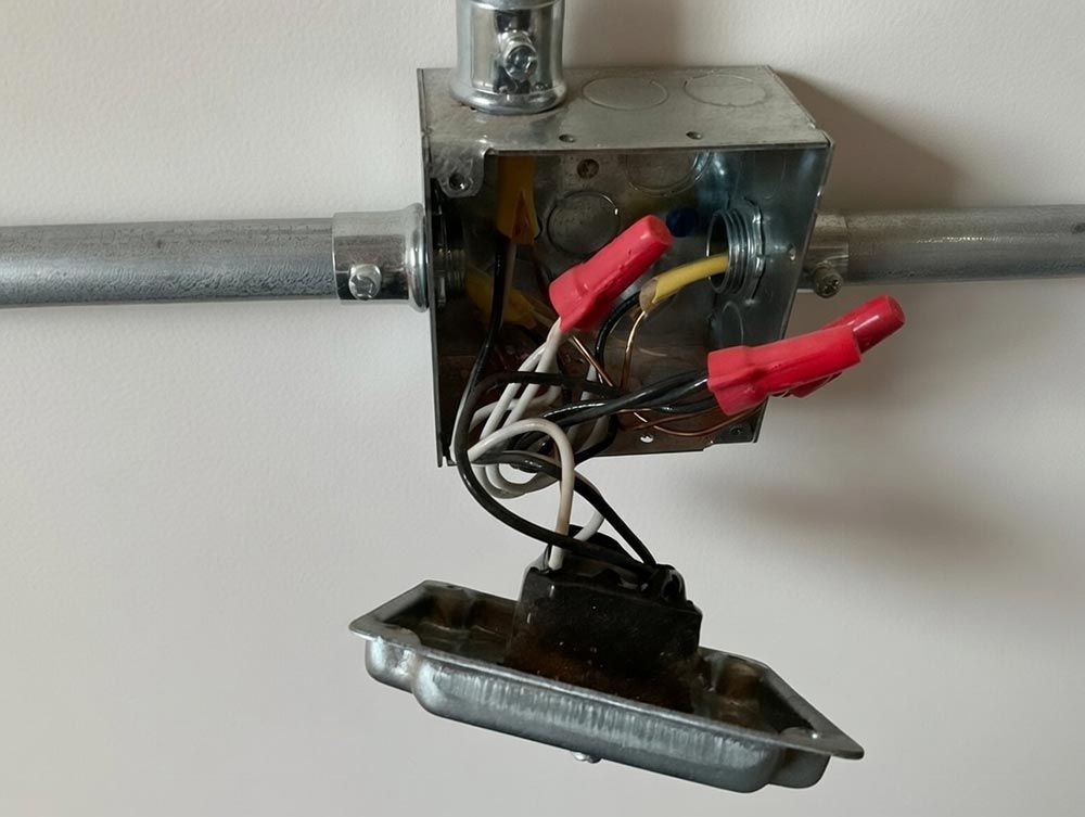 An open metal electrical junction box on a white wall with exposed wires connected by red wire nuts.