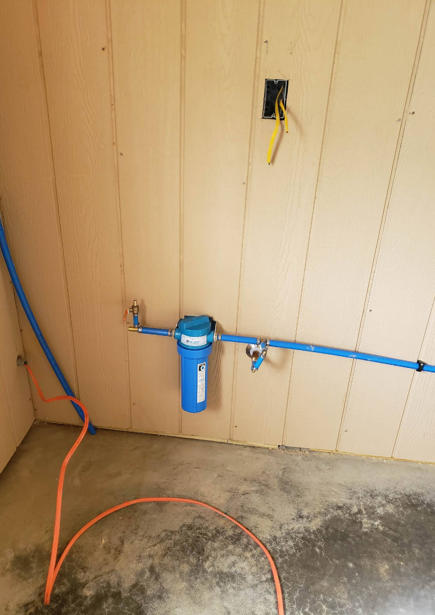 Water spigot with blue filter mounted on a tan wall, with hoses and concrete floor below.