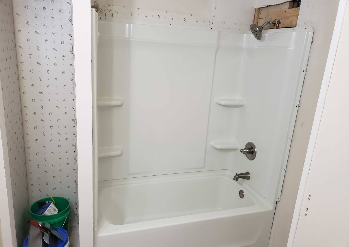 Small bathroom with a white bathtub and shower surround, grab bars, and a trash can beside it.