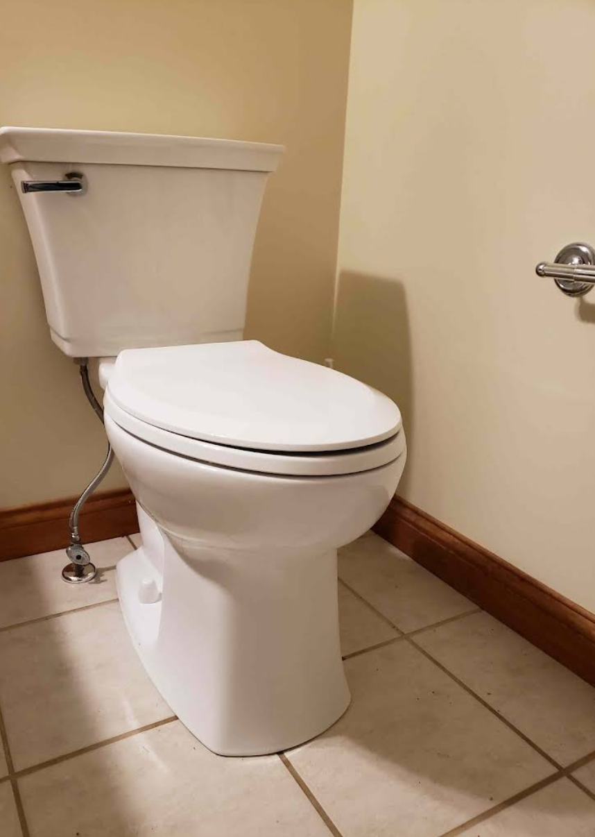 White toilet in a beige-tiled bathroom corner with a chrome flush handle and wooden baseboard.