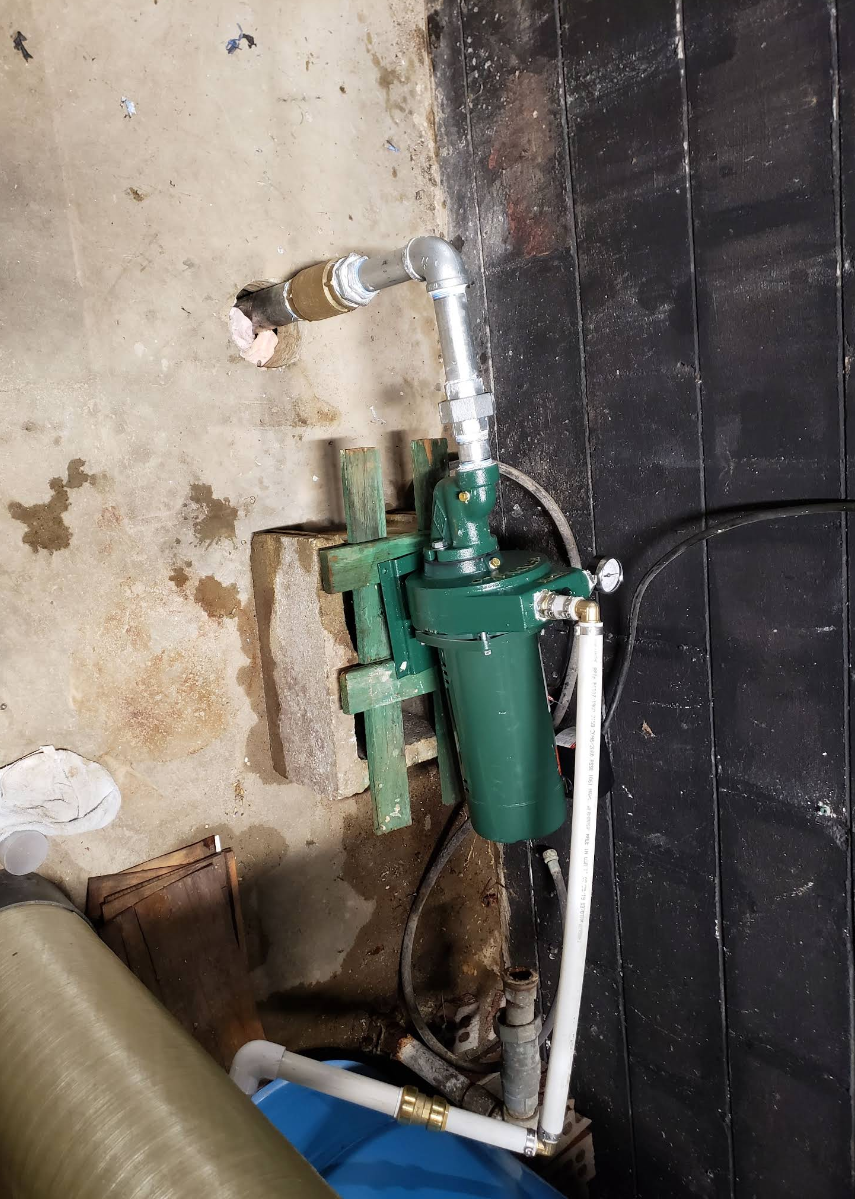 Green industrial pump and white pipes mounted against a concrete wall in a basement or utility area.