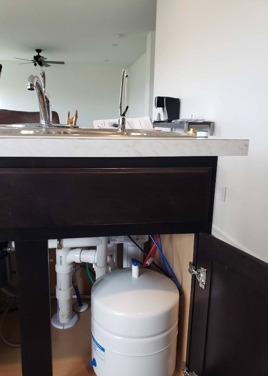 Open cabinet under bathroom sink showing white plumbing and a water filter tank under a black vanity.