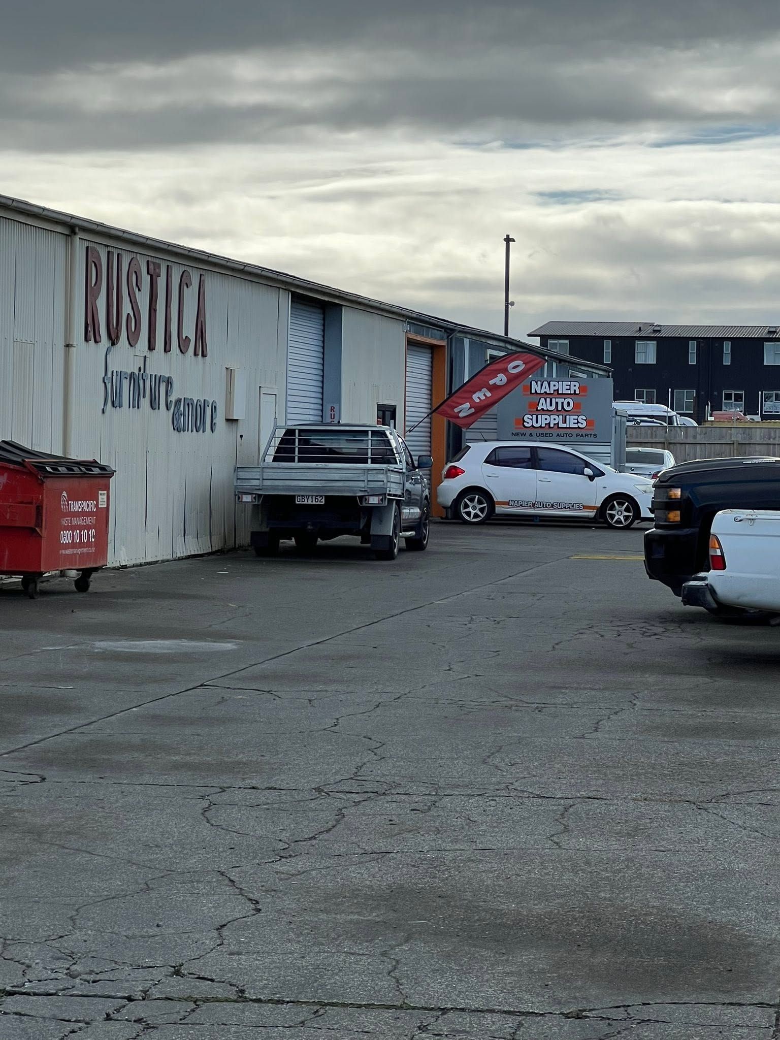 Snowy parking lot beside industrial buildings with parked cars and a red dumpster under cloudy skies