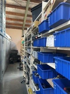 A narrow warehouse aisle with shelves of blue storage bins and stacked goods under a high ceiling.