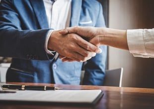 Two people shaking hands over a table with a document and pen. One person wears a blue suit.