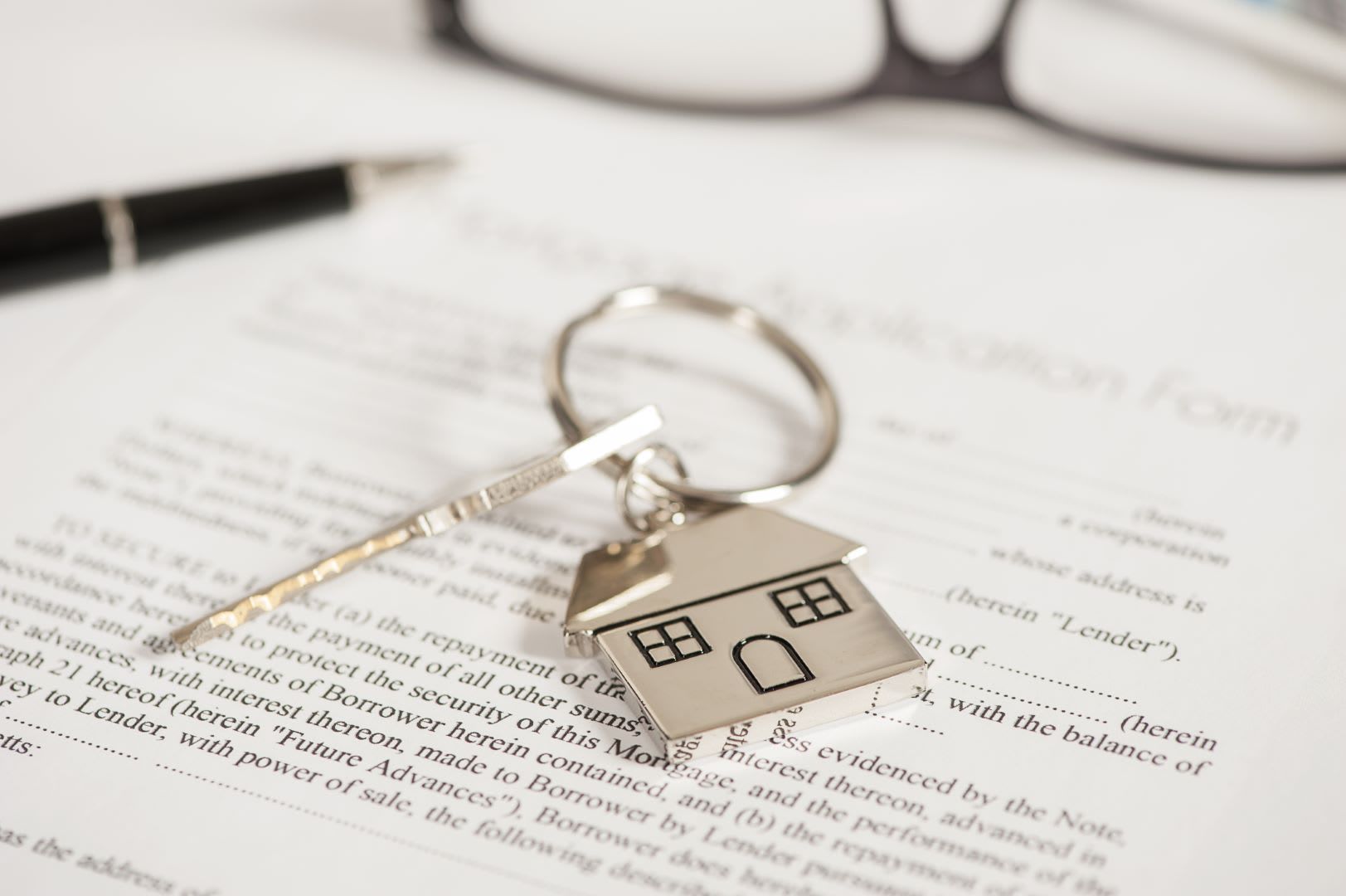 Keys with house-shaped tag on real estate document next to pen and glasses.