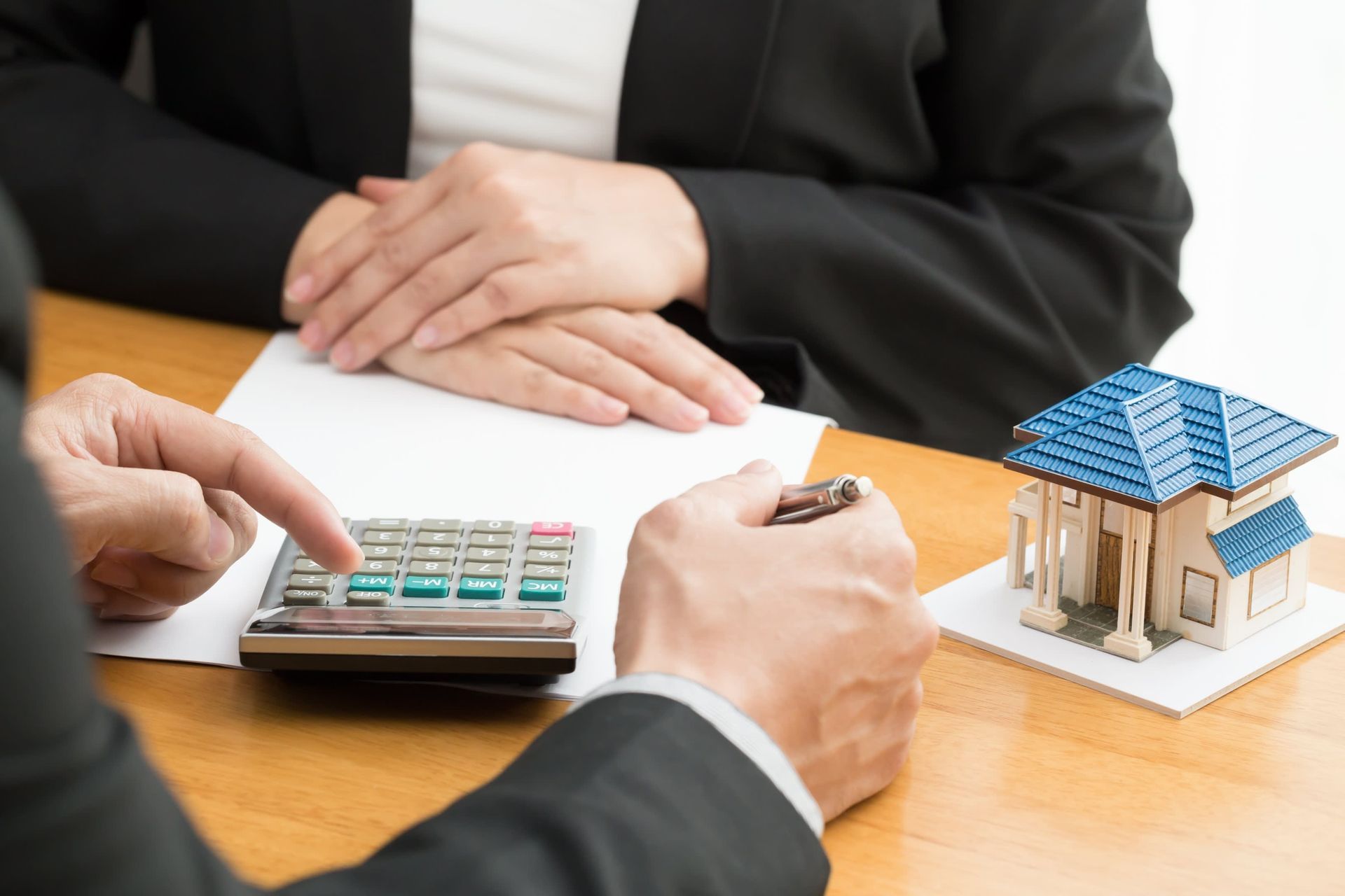 Person using a calculator with a miniature house on a desk; another person sits across from them, hands on a paper.