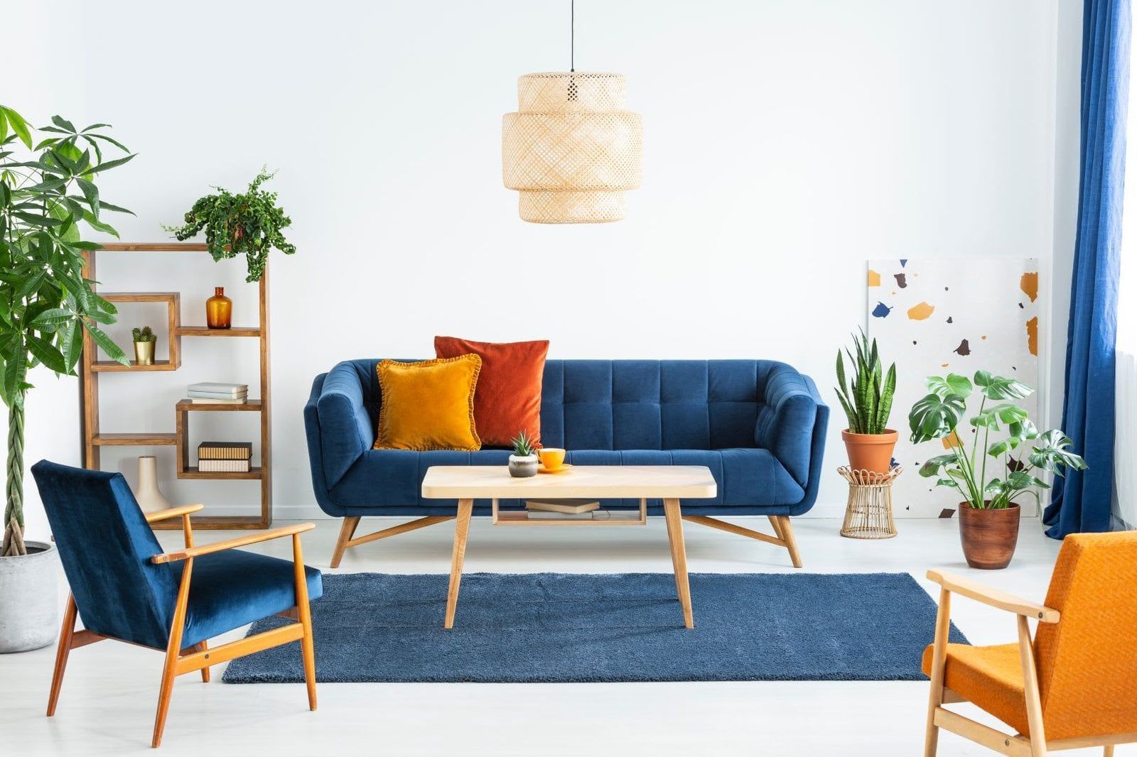 Living room with blue sofa, two armchairs, wood shelving, plants, and a woven pendant light.