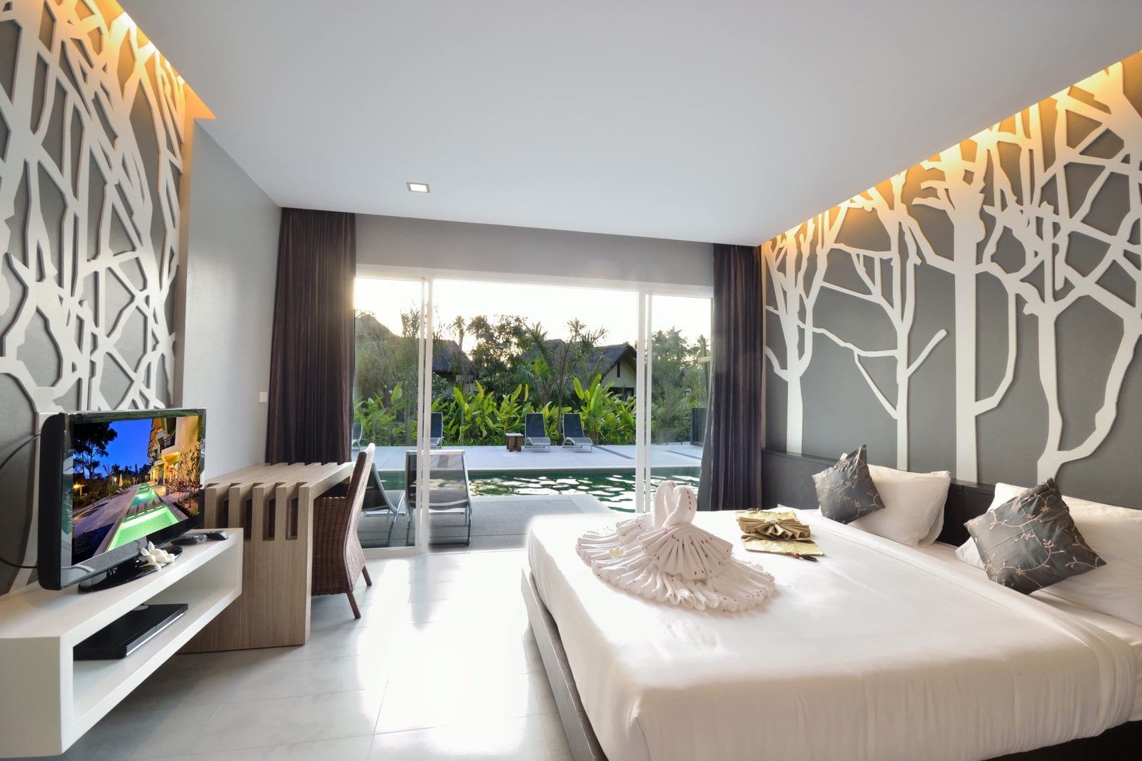 Hotel room with large bed, TV, and decorative tree designs on the walls overlooking a pool.