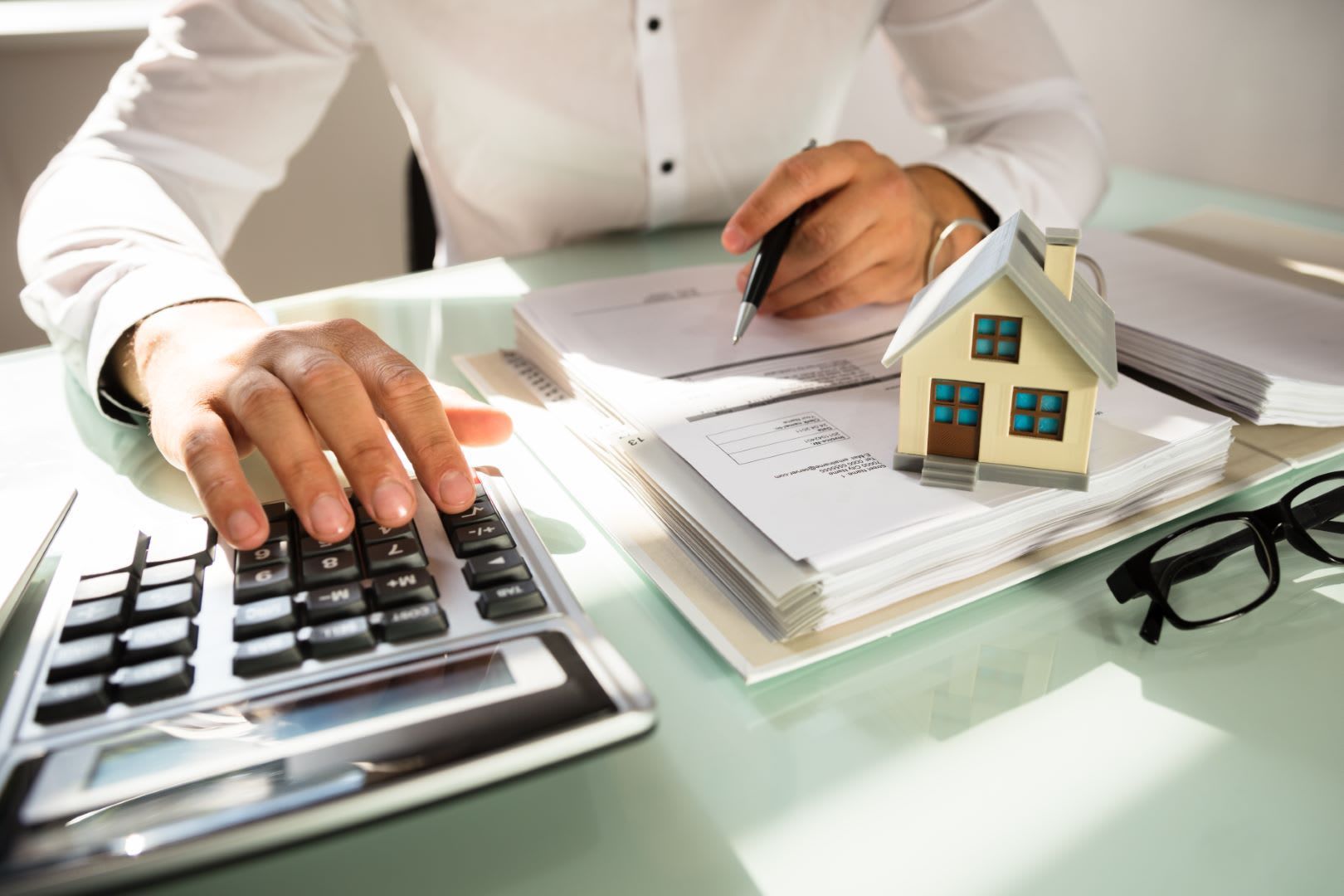 Person using a calculator, with paperwork, a small house model, and glasses on a desk.