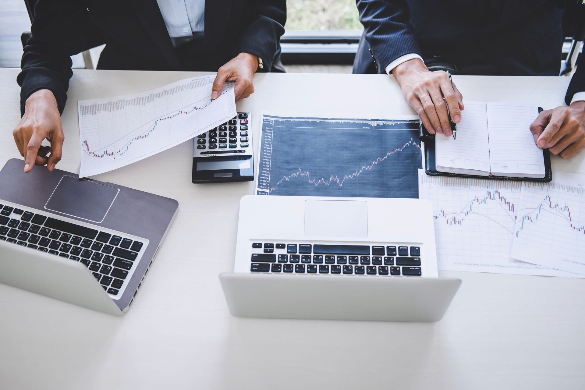 Two people in business attire reviewing charts and using laptops at a desk.