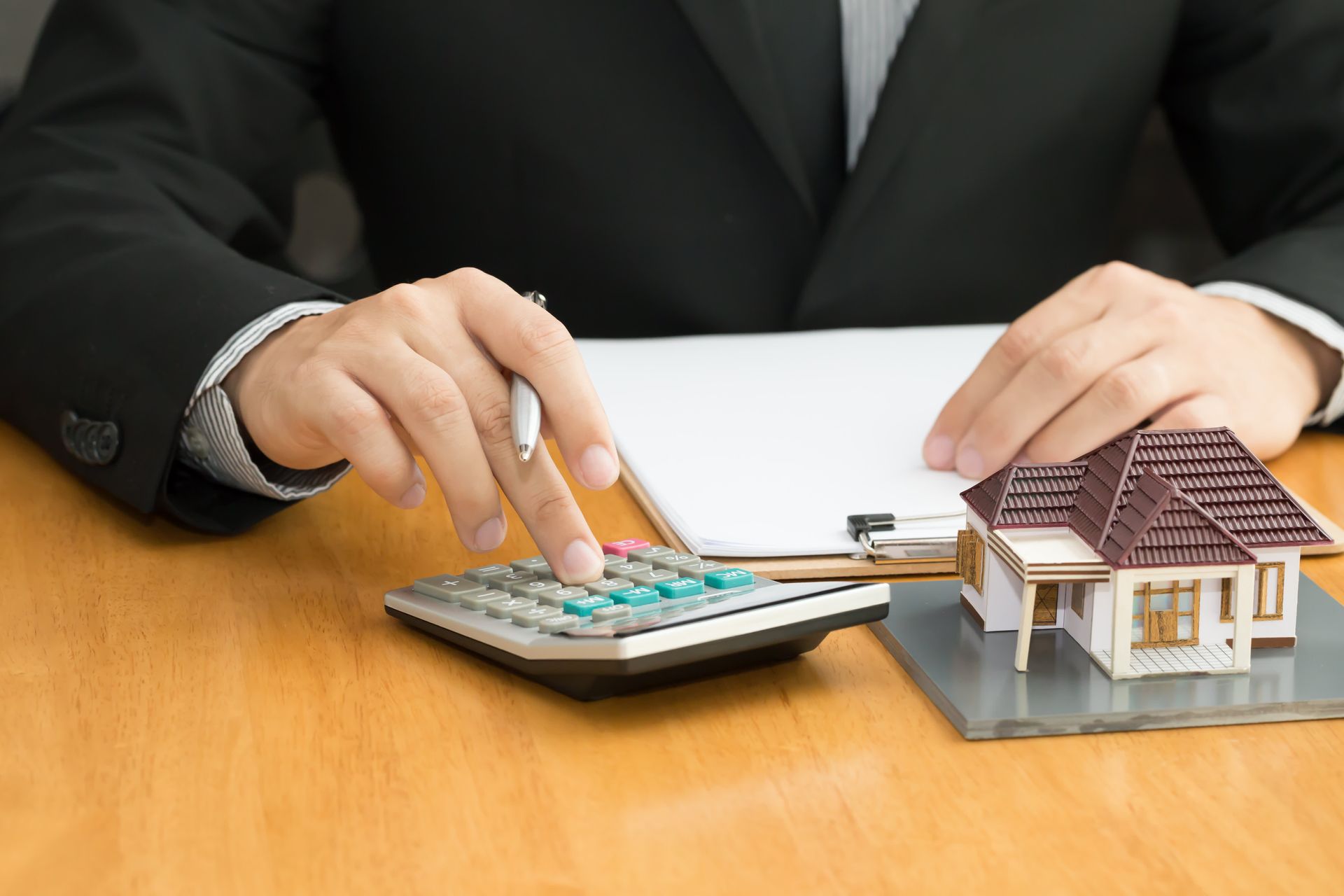 Person in suit calculating with a calculator next to a miniature house model. Clipboard present on a wooden desk.
