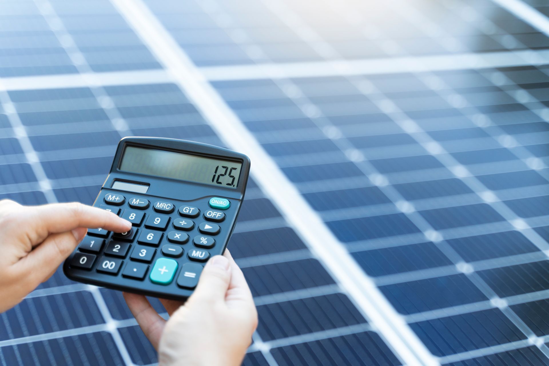 Hands using a calculator in front of solar panels, calculating energy costs.