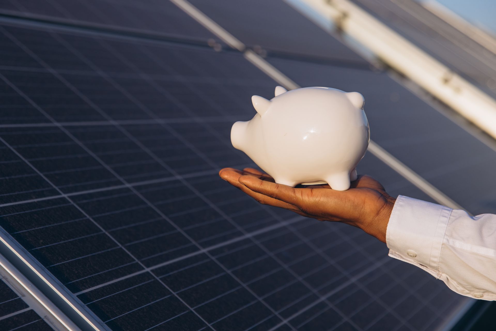 Hand holding a white piggy bank beside solar panels in sunlight