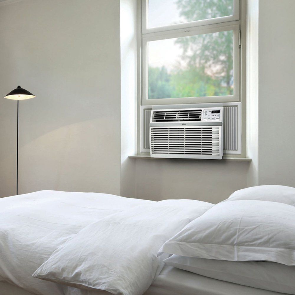 Habitación luminosa con ropa de cama blanca y un aire acondicionado de ventana sobre la cama.