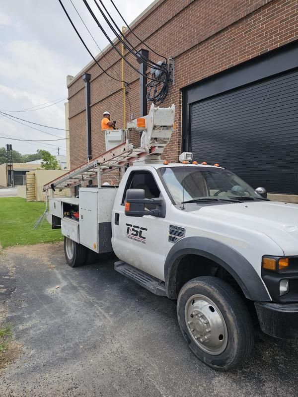 A utility truck with a worker in a raised bucket repairing overhead power lines near a brick building.