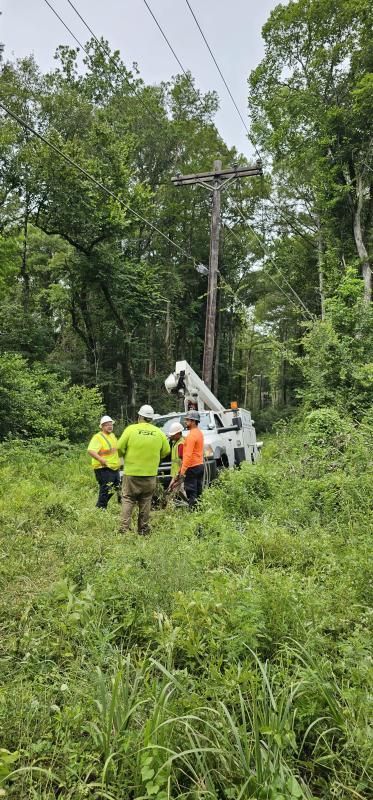 Linemen working on a utility pole in a wooded area.  A truck with a lift is present.