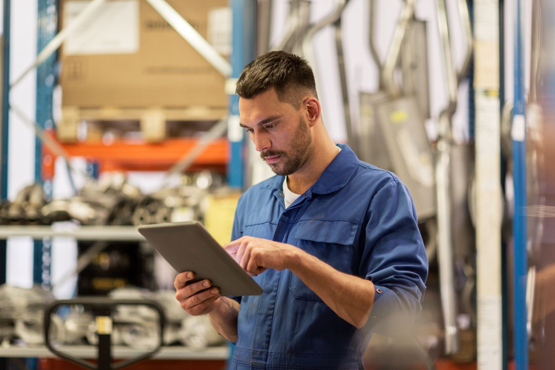 Man in blue work clothes using a tablet in a warehouse, surrounded by shelves and equipment.