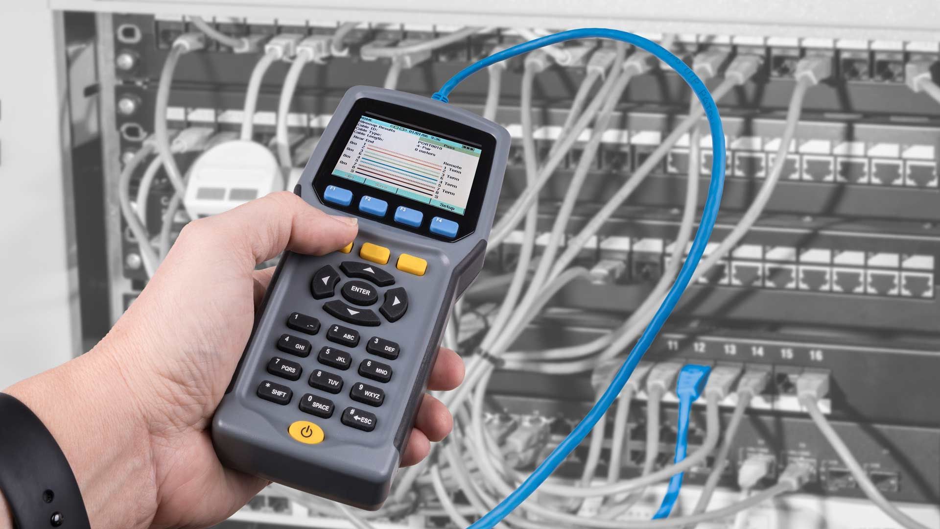 Hand holding a network cable tester, connected to a server rack with blue cables.