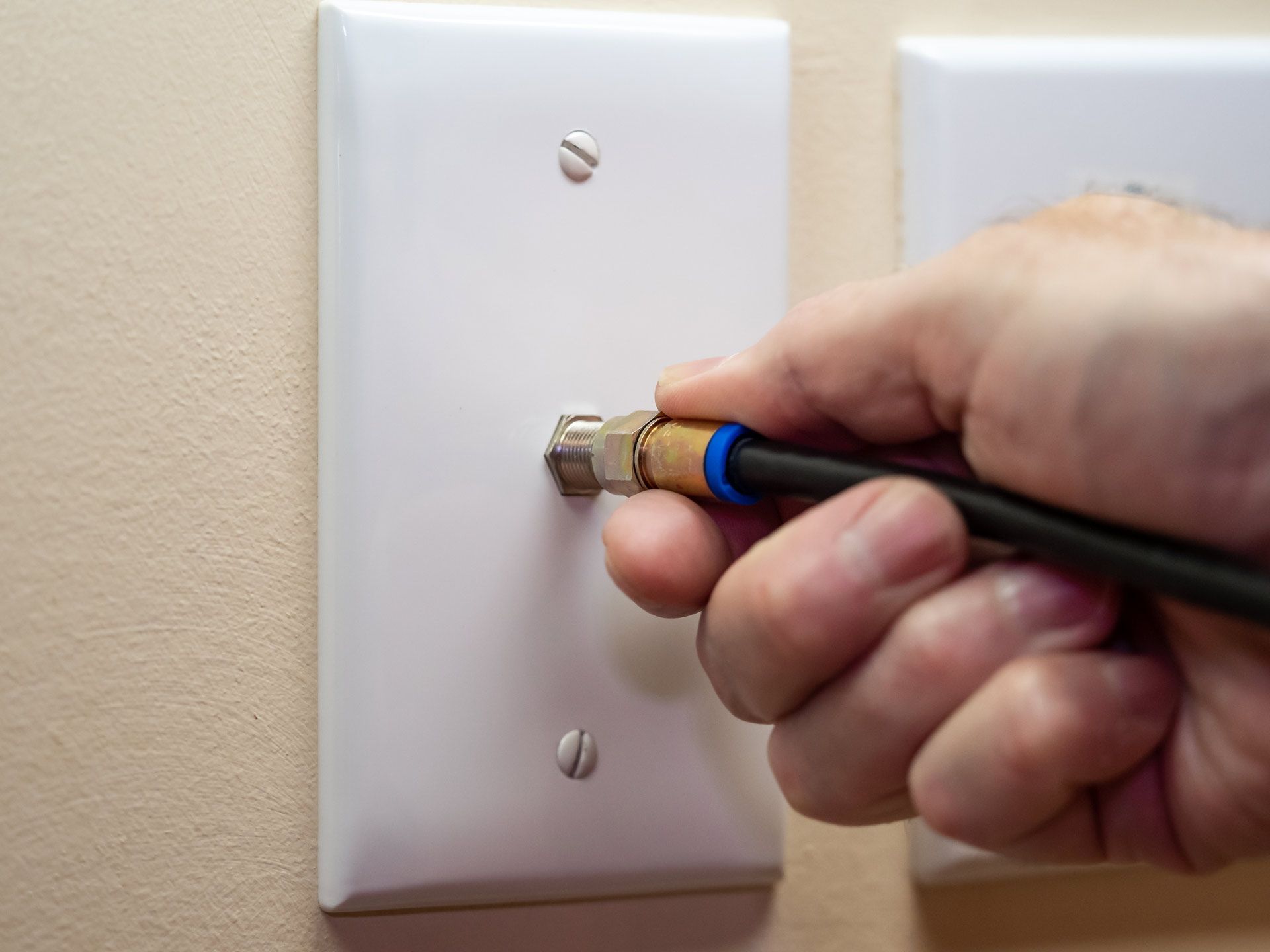 Hand plugging a coaxial cable into a wall outlet on a light-colored wall.