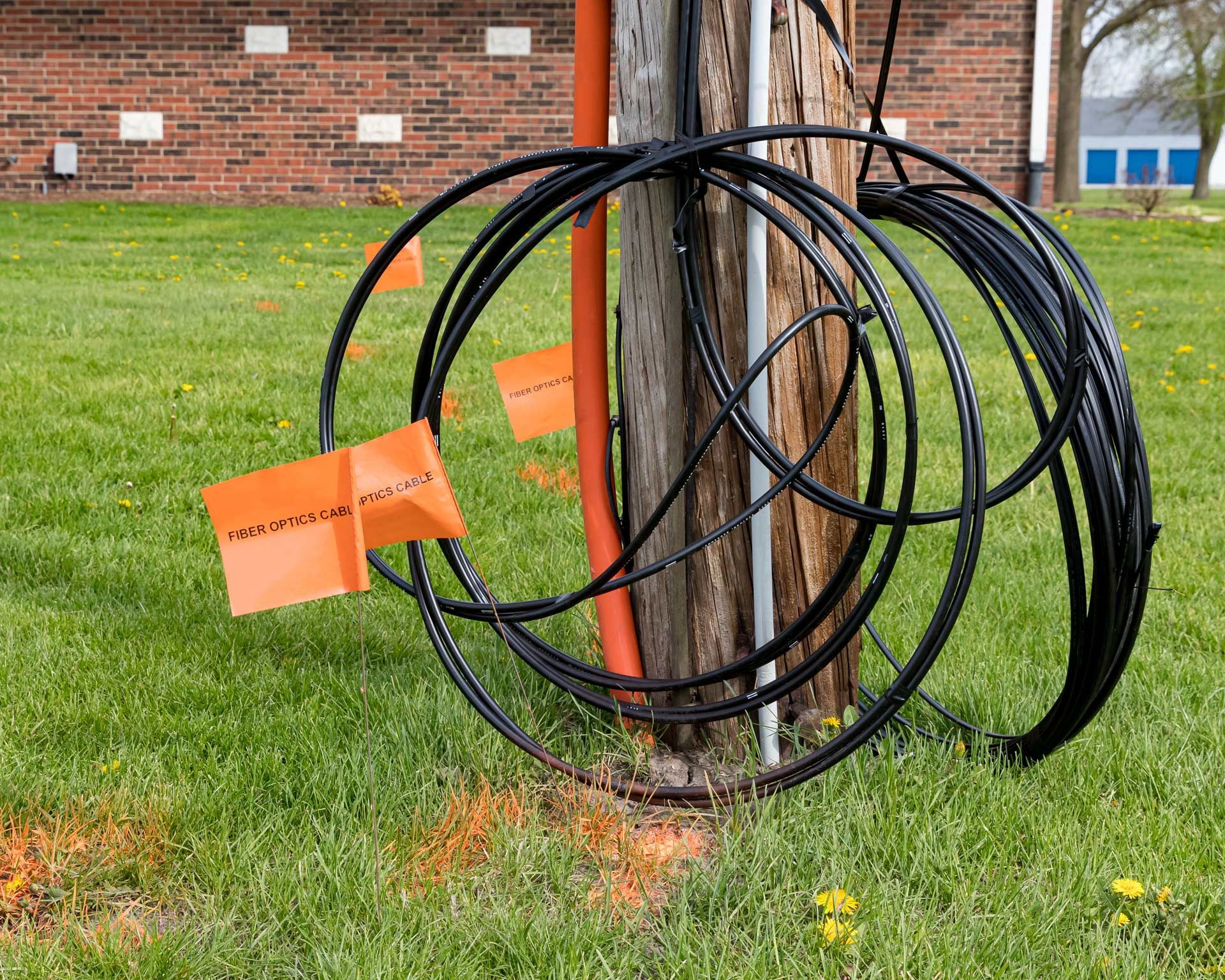 Black cable loops and orange markers around a utility pole in grass. Brick building in the background.