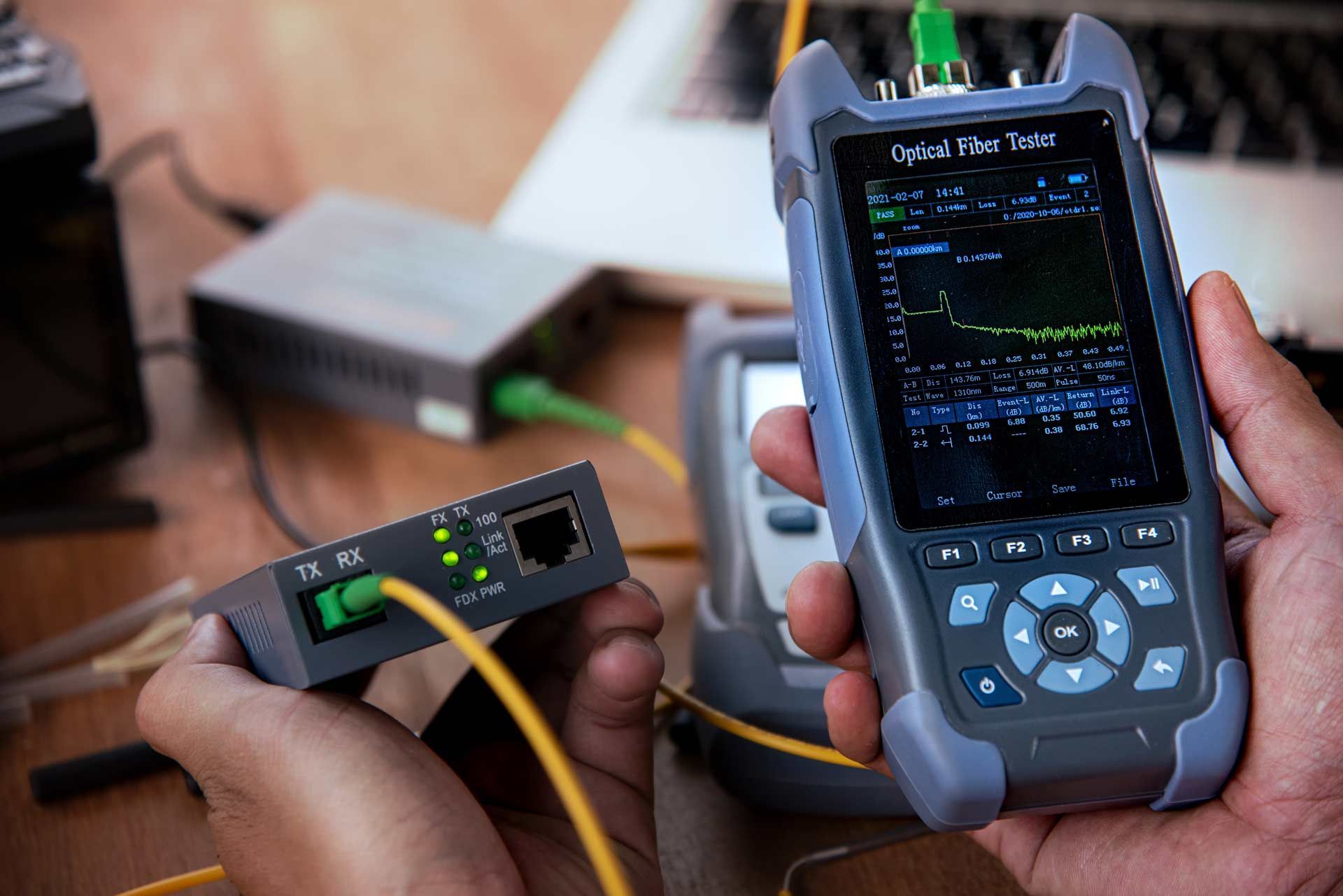 Hands holding an optical fiber tester, connected to a converter box and other equipment, testing network cables.