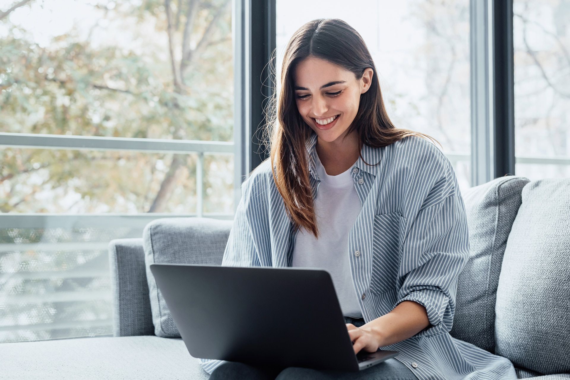 Woman smiling, using a laptop, sitting on a gray sofa near a large window.