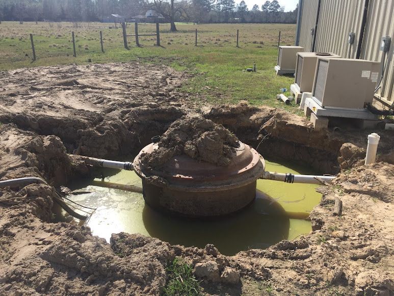 Rusted well head in a muddy pit beside a building, with yellow-green water around it.