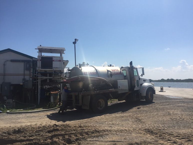Tank truck parked on a gravel shore beside water under a clear blue sky.