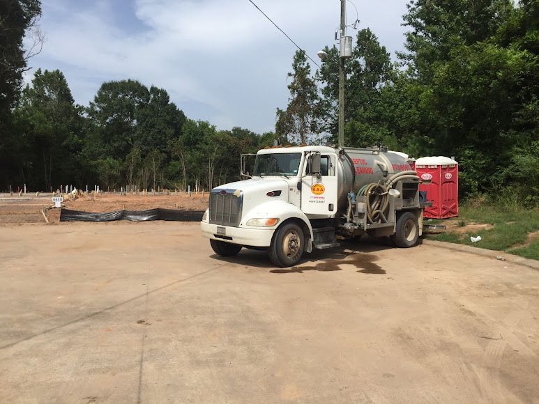 White tanker truck on a concrete lot near red portable toilets and trees under an overcast sky
