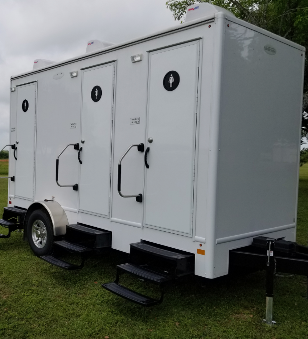 White portable restroom trailer with three doors and steps, parked on grass outdoors