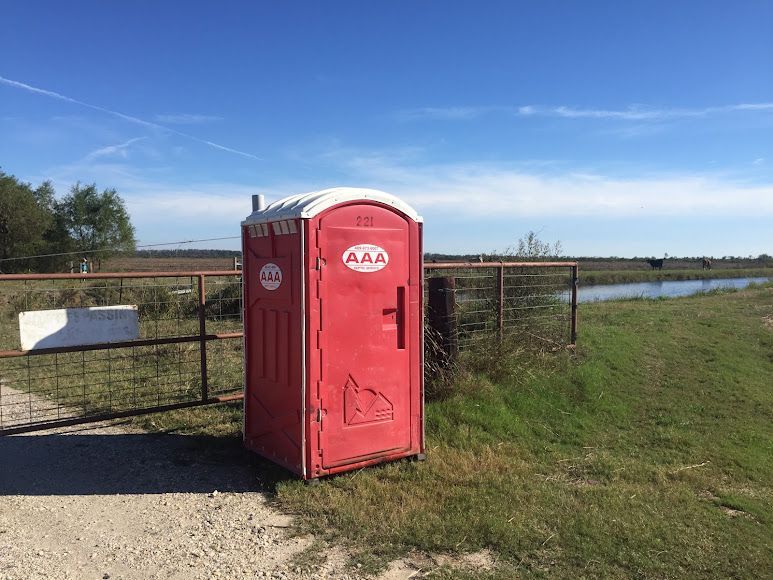 Red portable toilet beside a fence in a grassy field near water under a blue sky