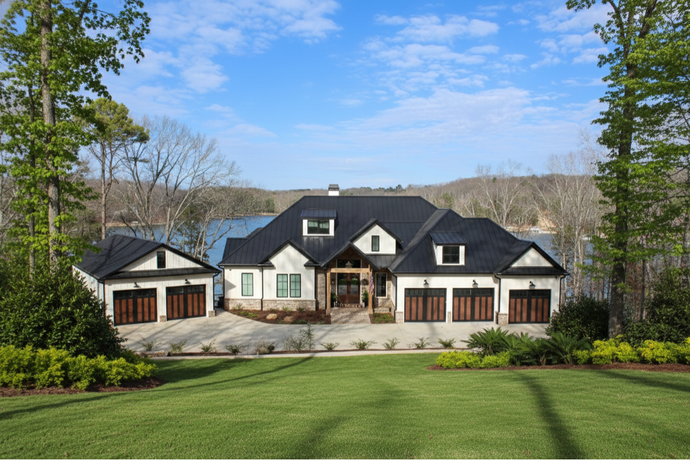 A large, white, multi-garage suburban home with a dark roof, nestled in a lush green, tree-filled landscape by a lake.